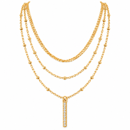 A gold multi-layer necklace featuring three distinct chain styles (curb, beaded, and delicate link) with a vertical bar pendant paved with clear stones, shown on a plain white background.