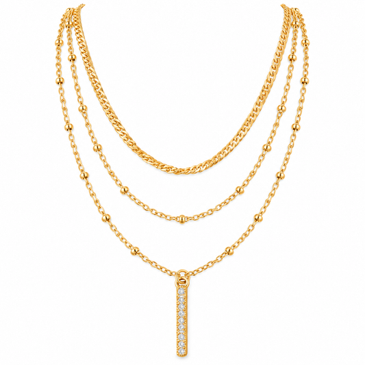 A gold multi-layer necklace featuring three distinct chain styles (curb, beaded, and delicate link) with a vertical bar pendant paved with clear stones, shown on a plain white background.