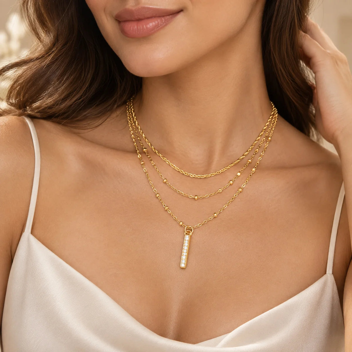 A close-up of a woman wearing a layered gold necklace with a vertical bar pendant set with small clear stones, paired with a white camisole.